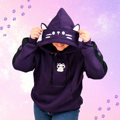 A person wearing a dark purple cat-themed hoodie with a hood shaped like a cat face, ears, and whiskers, holding the hood up over their head. The hoodie has a white cat face graphic on the hood and a small white cat graphic on the chest.